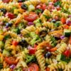 Rotini pasta salad with cherry tomatoes, cucumber, broccoli, zucchini, red and yellow bell peppers, shredded carrots, red onion, black olives, and parsley in a white serving bowl.