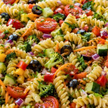Rotini pasta salad with cherry tomatoes, cucumber, broccoli, zucchini, red and yellow bell peppers, shredded carrots, red onion, black olives, and parsley in a white serving bowl.