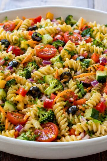 Rotini pasta salad with cherry tomatoes, cucumber, broccoli, zucchini, red and yellow bell peppers, shredded carrots, red onion, black olives, and parsley in a white serving bowl.