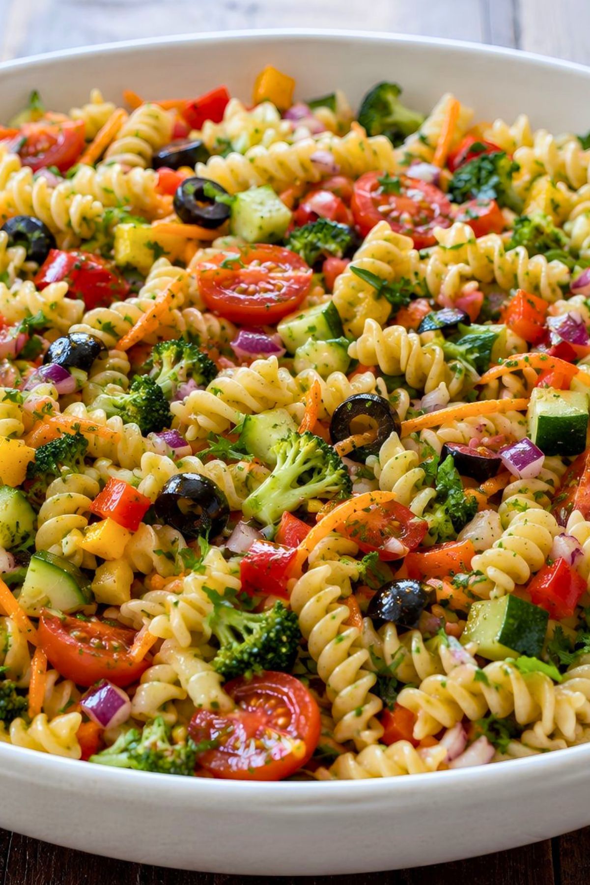 Rotini pasta salad with cherry tomatoes, cucumber, broccoli, zucchini, red and yellow bell peppers, shredded carrots, red onion, black olives, and parsley in a white serving bowl.