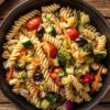 Garden vegetable pasta salad with rotini, cherry tomatoes, broccoli, zucchini, cucumber, carrots, red onion, and black olives in a black bowl.