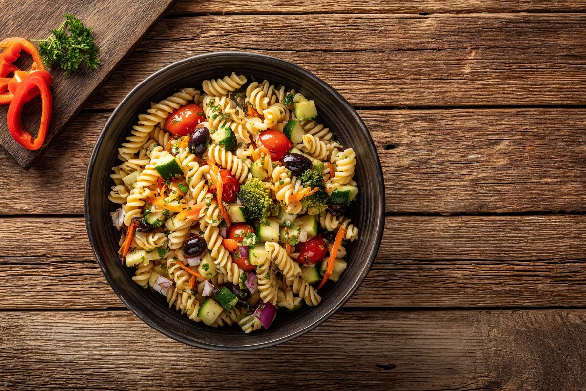 Easy garden vegetable pasta salad with rotini, fresh vegetables, cherry tomatoes, broccoli, and black olives on a rustic wooden table.