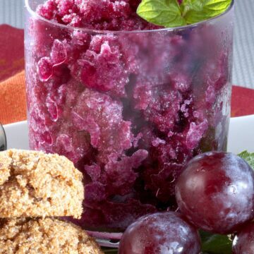 Grape granita served in a clear glass with icy purple crystals, fresh mint, red grapes, and oat cookies on a white plate. A bright frozen dessert photo with a close-up view of the fruity texture.