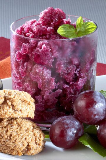 Grape granita served in a clear glass with icy purple crystals, fresh mint, red grapes, and oat cookies on a white plate. A bright frozen dessert photo with a close-up view of the fruity texture.