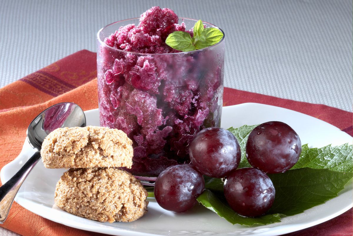 Grape granita in a clear glass topped with mint, served on a white plate with red grapes, green leaves, oat cookies, and a spoon. A bright frozen grape dessert with icy purple texture.