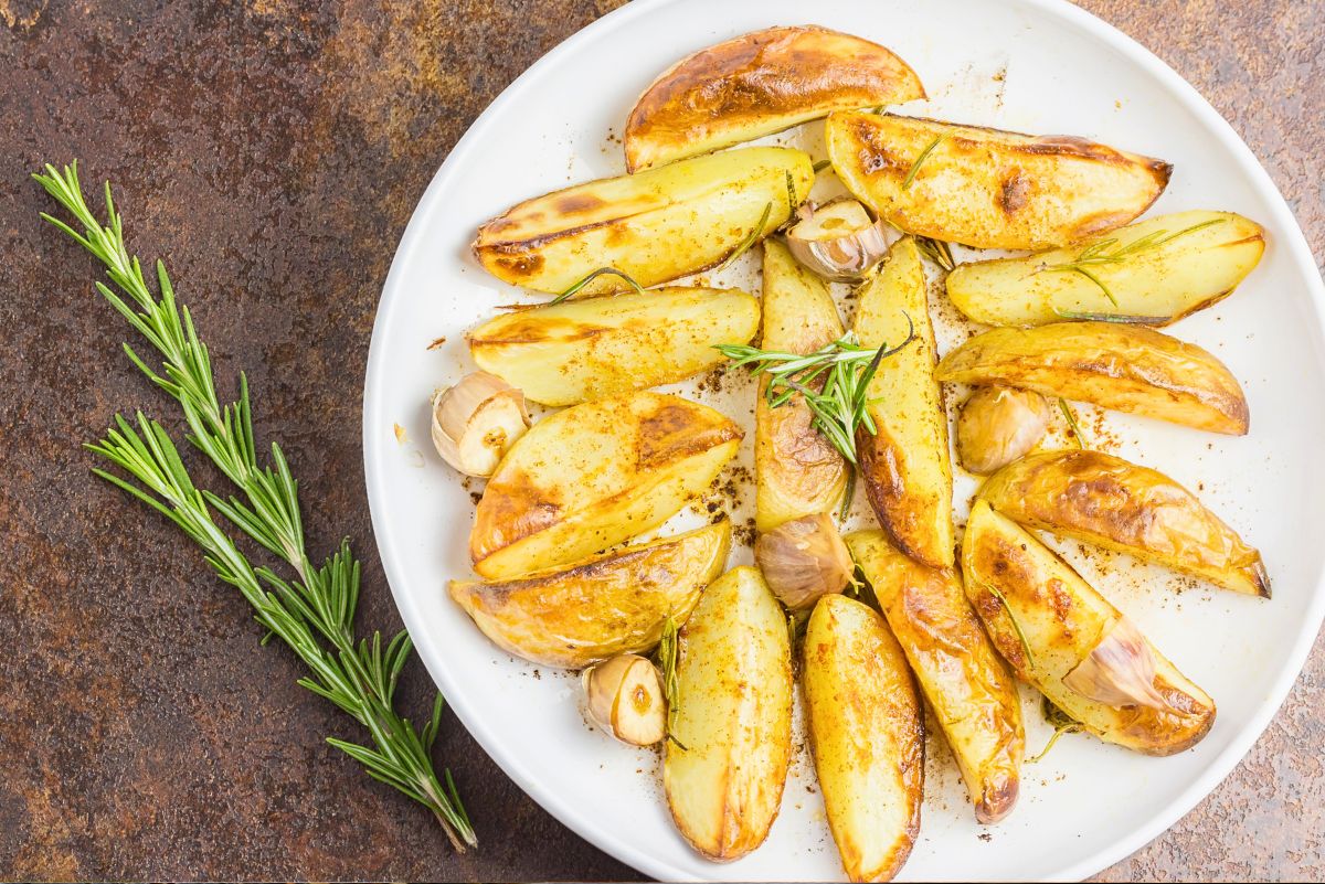 Grilled fingerling potatoes with roasted garlic cloves, rosemary, and southwest seasoning on a white plate, with fresh rosemary sprigs on a rustic brown surface.