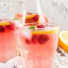 Close-up of pink raspberry lemonade vodka cooler drinks with ice, raspberries, lemon slices, and mint, with a pitcher in the background and fresh lemon on the side.