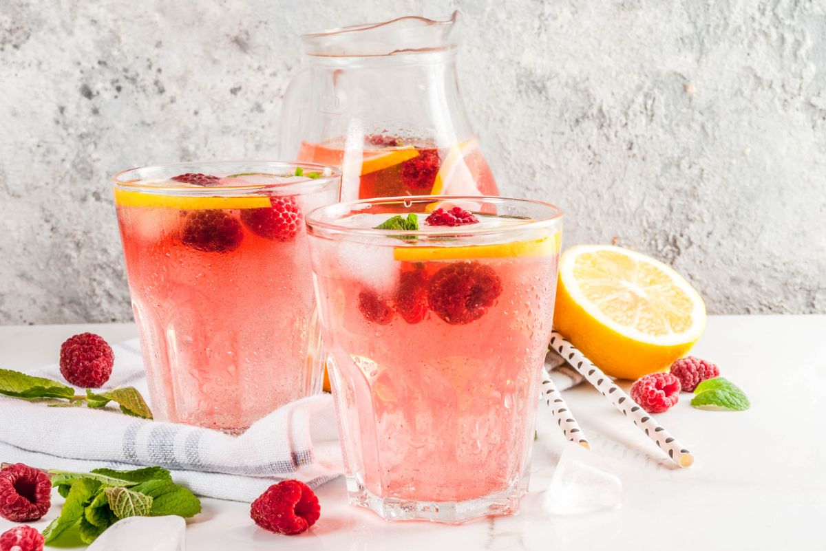 Pink raspberry lemonade vodka cooler drinks in clear glasses with ice, lemon slices, raspberries, and mint, served beside a glass pitcher, fresh lemon, paper straws, and scattered raspberries.