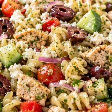 Greek chicken pasta salad in a white bowl with grilled chicken, rotini pasta, cucumber, cherry tomatoes, kalamata olives, red onion, feta cheese, and chopped herbs.