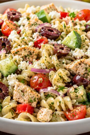 Greek chicken pasta salad in a white bowl with grilled chicken, rotini pasta, cucumber, cherry tomatoes, kalamata olives, red onion, feta cheese, and chopped herbs.