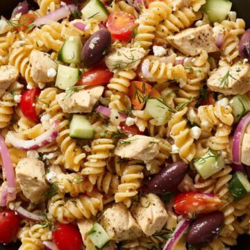 Close-up of Greek chicken pasta salad with rotini, cucumbers, tomatoes, Kalamata olives, red onion, feta, and dill in a black bowl.