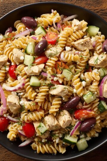 Close-up of Greek chicken pasta salad with rotini, cucumbers, tomatoes, Kalamata olives, red onion, feta, and dill in a black bowl.