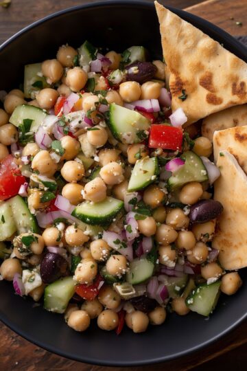 Close-up of Greek chickpea salad with cucumbers, tomatoes, red onion, olives, feta, and crispy garlic pita chips.