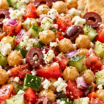 Greek chickpea salad with cucumbers, tomatoes, red onion, kalamata olives, feta cheese, herbs, and garlic pita chips in a shallow bowl.