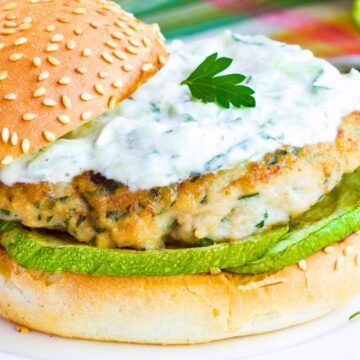 Greek style chicken zucchini burger on a sesame bun with grilled zucchini, lettuce, creamy tzatziki sauce, and a parsley garnish on top.