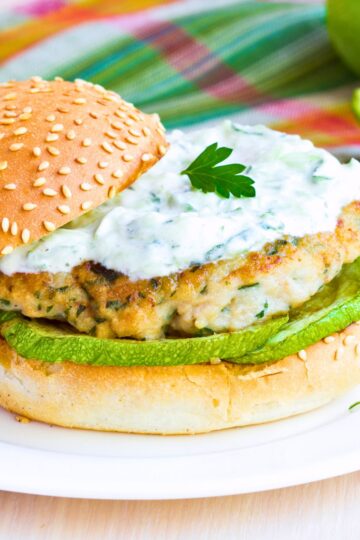 Greek style chicken zucchini burger on a sesame bun with grilled zucchini, lettuce, creamy tzatziki sauce, and a parsley garnish on top.