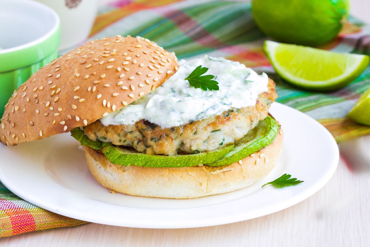 Greek style chicken zucchini burger on a sesame bun with grilled zucchini, creamy tzatziki sauce, parsley garnish, and lime wedges on a white plate.