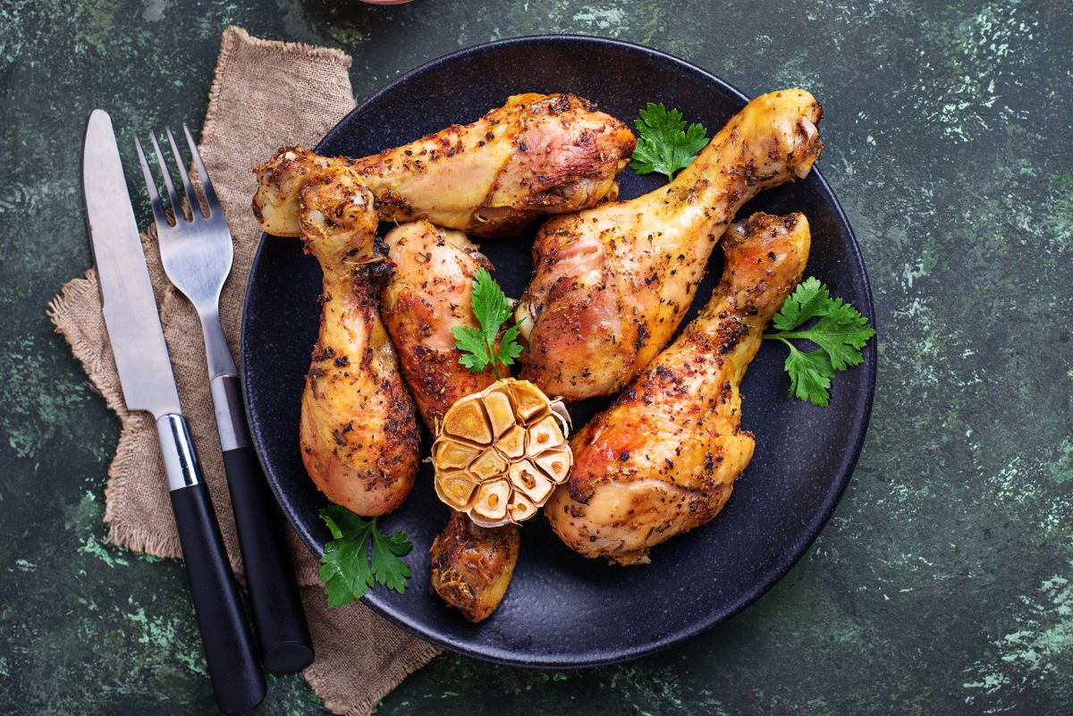 Grilled chicken drumsticks with maple beer glaze on a black plate, topped with roasted garlic and fresh parsley. A fork and knife sit on burlap beside the plate on a dark green textured surface.