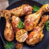 Grilled chicken drumsticks with a maple beer glaze on a black plate, garnished with roasted garlic and fresh parsley. A small bowl of sauce sits nearby on a dark green surface.