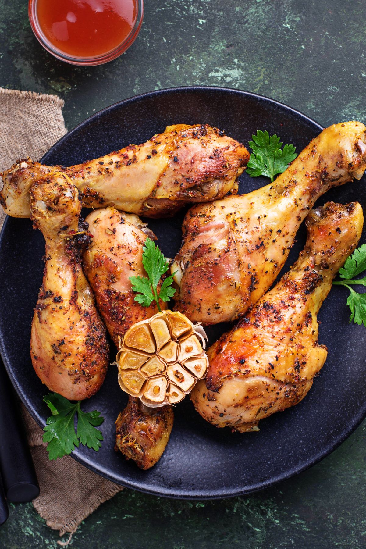 Grilled chicken drumsticks with a maple beer glaze on a black plate, garnished with roasted garlic and fresh parsley. A small bowl of sauce sits nearby on a dark green surface.