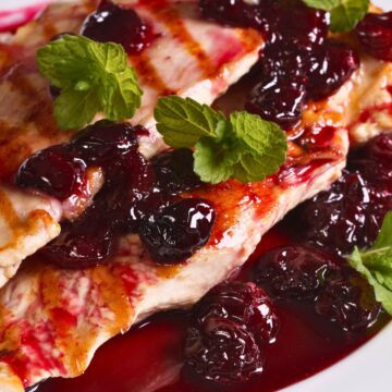 Grilled chicken breast topped with smoky cherry sauce, whole cherries, and fresh mint on a white plate.