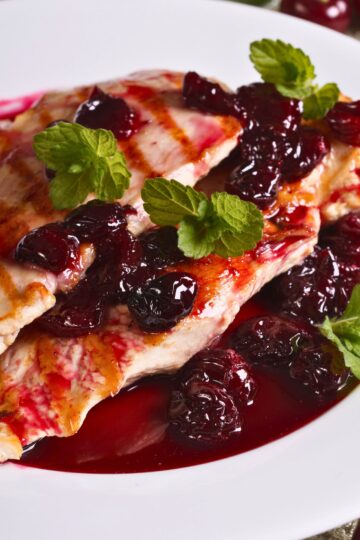 Grilled chicken breast topped with smoky cherry sauce, whole cherries, and fresh mint on a white plate.