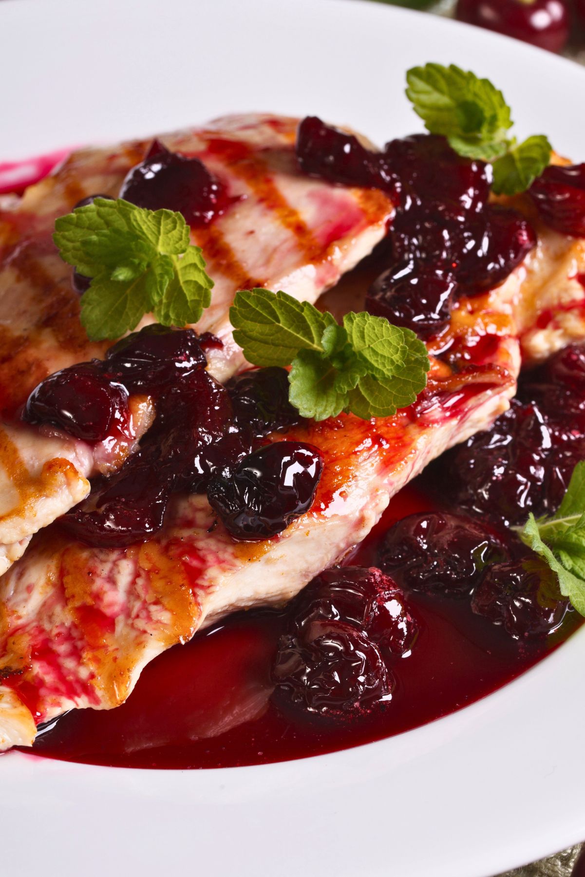 Grilled chicken breast topped with smoky cherry sauce, whole cherries, and fresh mint on a white plate.