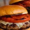 Grilled pepperoni pizza burger on a toasted bun with a beef patty, melted cheese, grilled tomato slices, and turkey pepperoni, with a second burger blurred in the background.