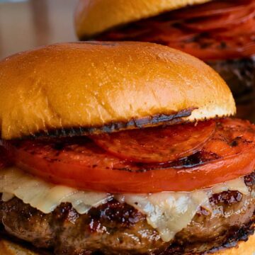 Grilled pepperoni pizza burger on a toasted bun with a beef patty, melted cheese, grilled tomato slices, and turkey pepperoni, with a second burger blurred in the background.