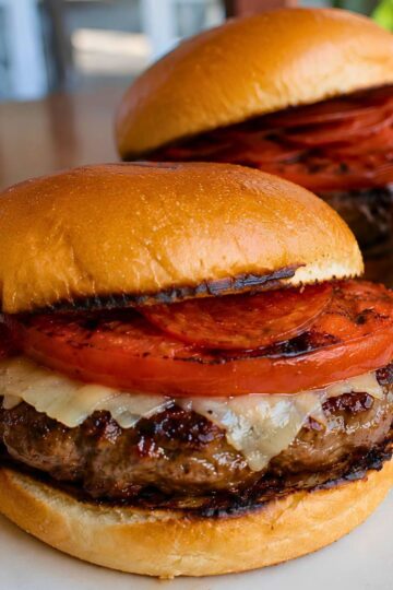 Grilled pepperoni pizza burger on a toasted bun with a beef patty, melted cheese, grilled tomato slices, and turkey pepperoni, with a second burger blurred in the background.