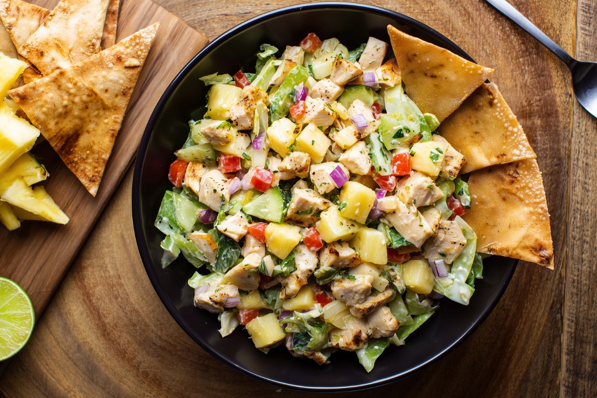 Hawaiian chicken salad with chopped pineapple, cucumbers, lettuce, red peppers, red onion, and crispy chips in a black bowl.