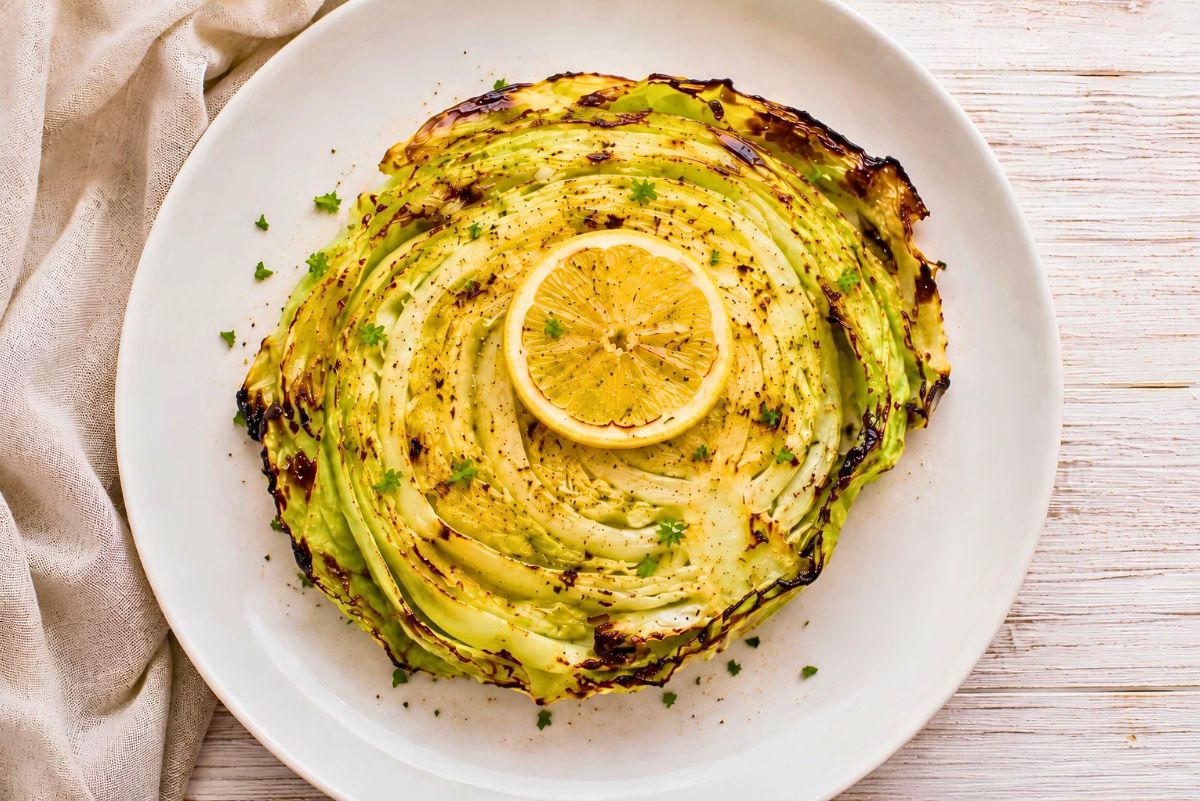 Grilled cabbage steak with charred edges, black pepper, herbs, and a fresh lemon slice on a white plate. A simple 30 minute cookout side dish served on a whitewashed wood table.