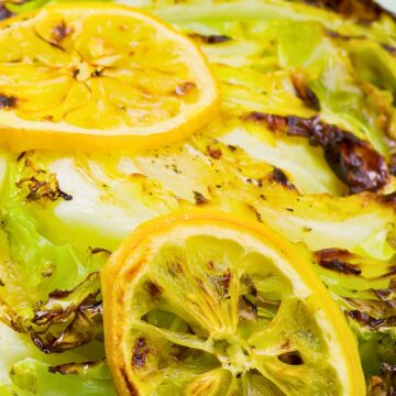 Grilled cabbage steaks with charred edges, black pepper, and roasted lemon slices on a turquoise plate. A bright summer cookout side dish with fresh lemon and lightly browned cabbage.