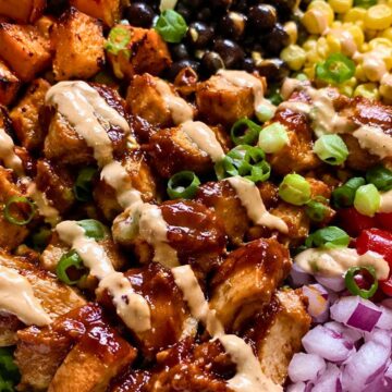 High protein BBQ chicken sweet potato bowl with black beans, corn, roasted sweet potatoes, red onion, lettuce, green onions, and creamy sauce drizzle.