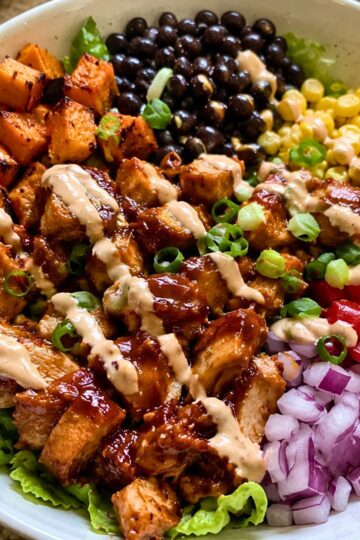 High protein BBQ chicken sweet potato bowl with black beans, corn, roasted sweet potatoes, red onion, lettuce, green onions, and creamy sauce drizzle.