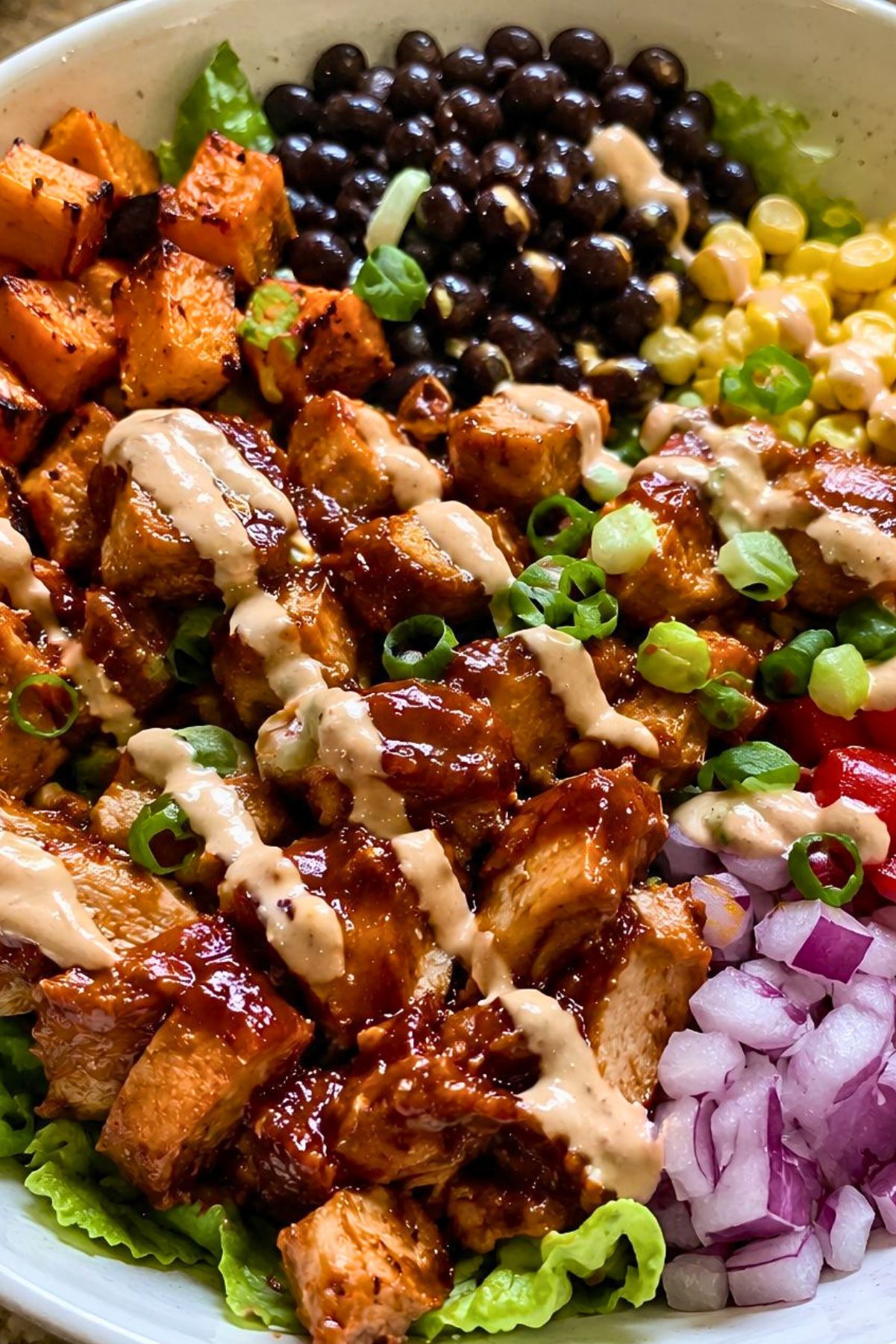 High protein BBQ chicken sweet potato bowl with black beans, corn, roasted sweet potatoes, red onion, lettuce, green onions, and creamy sauce drizzle.