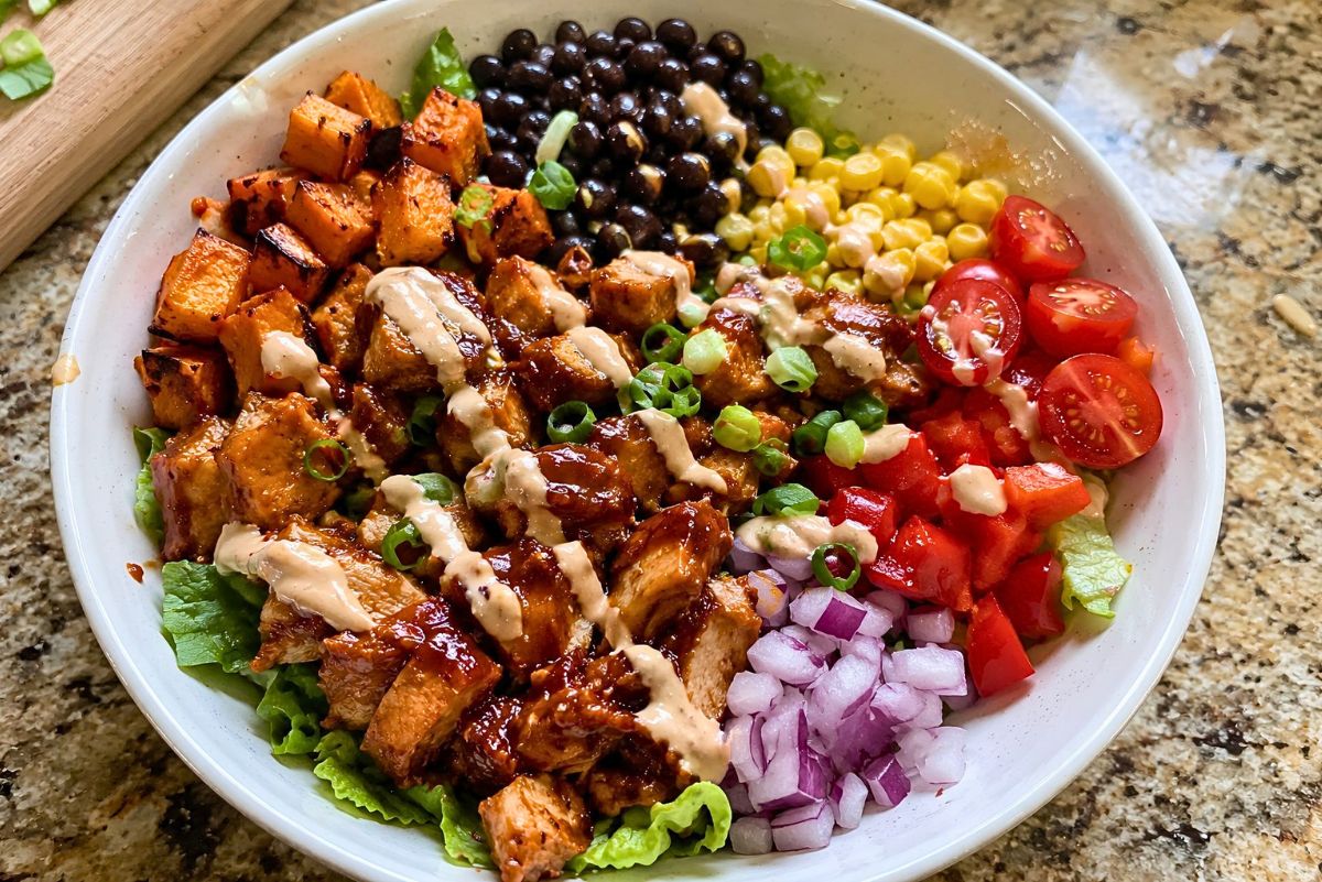 High protein BBQ chicken sweet potato bowl with roasted sweet potatoes, black beans, corn, cherry tomatoes, red onion, lettuce, green onions, and creamy sauce drizzle in a white bowl.