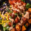 Close-up of a BBQ chicken sweet potato protein bowl with roasted sweet potatoes, black beans, corn, tomatoes, red onion, lettuce, and creamy dressing.