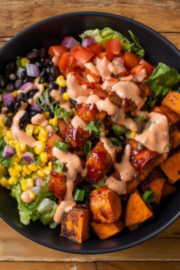 Close-up of a BBQ chicken sweet potato protein bowl with roasted sweet potatoes, black beans, corn, tomatoes, red onion, lettuce, and creamy dressing.