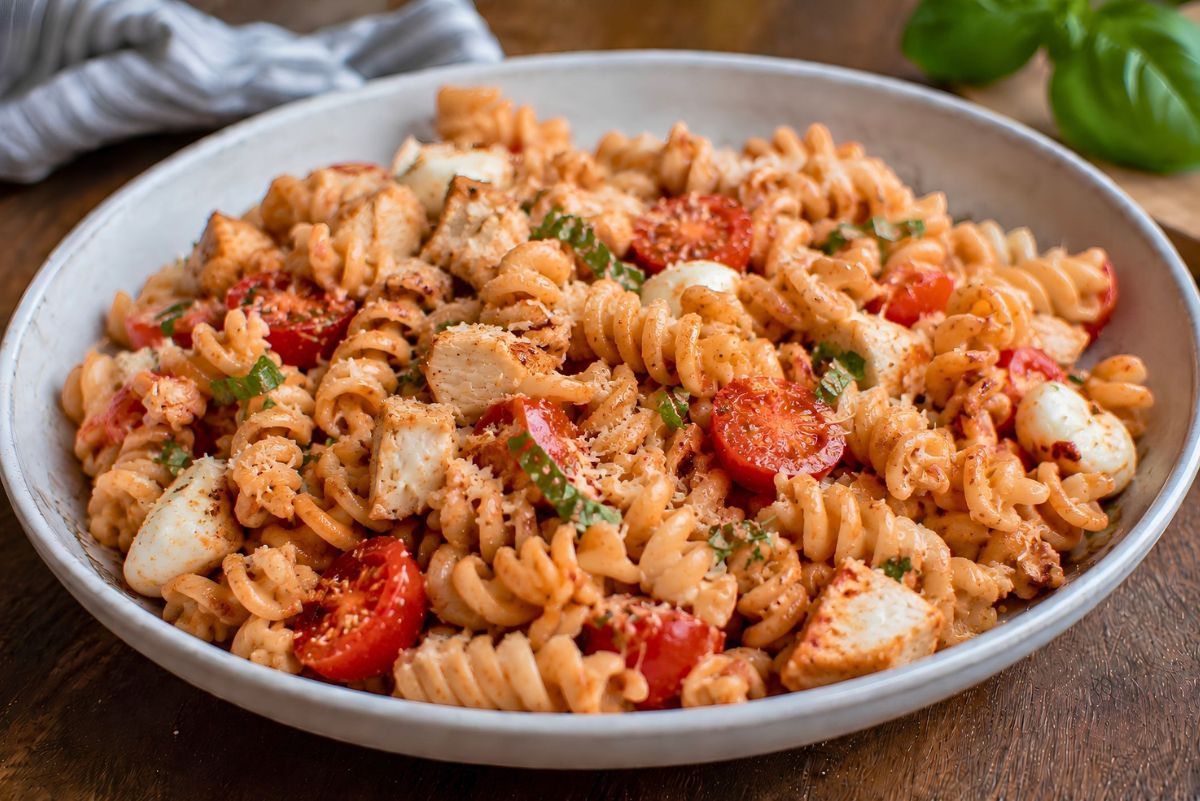 High protein chicken Parmesan pasta salad with rotini, grilled chicken, cherry tomatoes, mozzarella pearls, basil, marinara sauce, and grated Parmesan in a shallow white bowl.