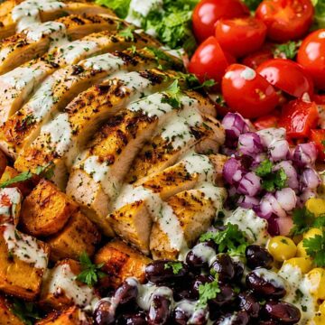 High protein chicken sweet potato bowl with sliced grilled chicken, roasted sweet potatoes, black beans, corn, tomatoes, red onion, romaine lettuce, cilantro, and creamy herb dressing in a large ceramic bowl.