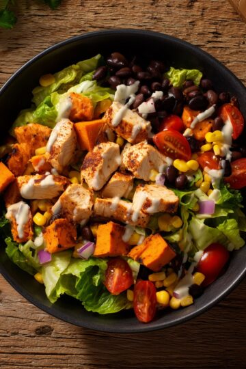High-protein chicken sweet potato bowl with roasted sweet potatoes, seasoned chicken, black beans, corn, cherry tomatoes, lettuce, red onion, and creamy dressing.