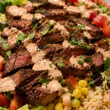 Steak burrito bowl with sliced grilled steak, brown rice, corn, black beans, chopped tomatoes, lettuce, cilantro, and creamy sauce in a shallow bowl.