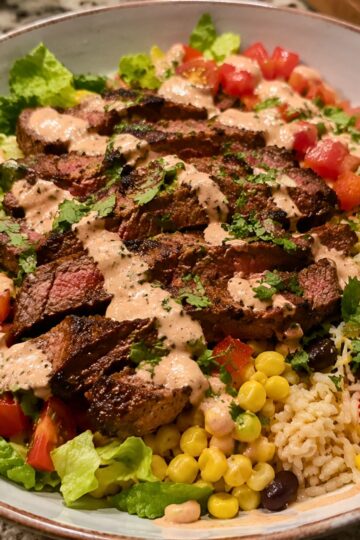 Steak burrito bowl with sliced grilled steak, brown rice, corn, black beans, chopped tomatoes, lettuce, cilantro, and creamy sauce in a shallow bowl.