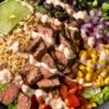 Overhead view of a high protein steak burrito bowl with sliced medium-cooked steak, brown rice, black beans, corn, chopped tomatoes, red onion, romaine lettuce, lime wedges, and creamy chipotle-style sauce in a black bowl.
