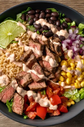 Overhead view of a high protein steak burrito bowl with sliced medium-cooked steak, brown rice, black beans, corn, chopped tomatoes, red onion, romaine lettuce, lime wedges, and creamy chipotle-style sauce in a black bowl.