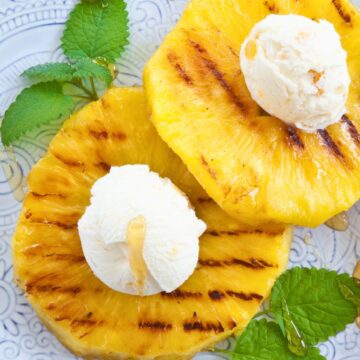 Honey glazed grilled pineapple rings topped with vanilla ice cream, fresh mint leaves, and drizzled honey on a blue patterned plate.