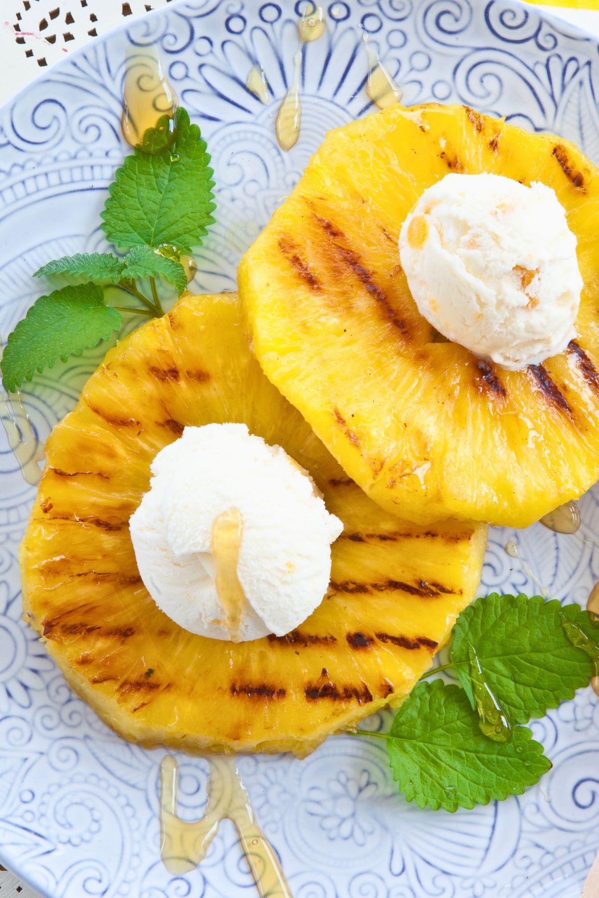 Honey glazed grilled pineapple rings topped with vanilla ice cream, fresh mint leaves, and drizzled honey on a blue patterned plate.