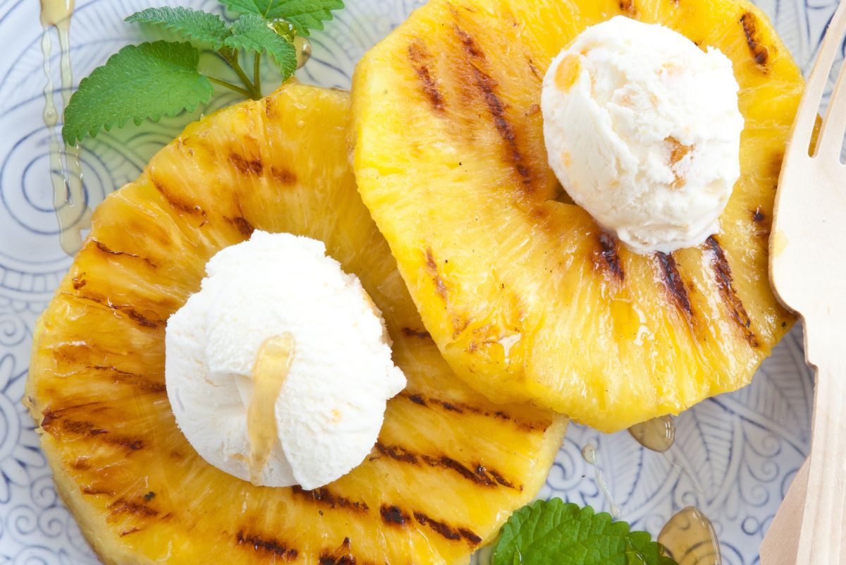 Honey glazed grilled pineapple rings topped with vanilla ice cream and fresh mint on a blue patterned plate, with a drizzle of honey and a wooden serving utensil nearby.