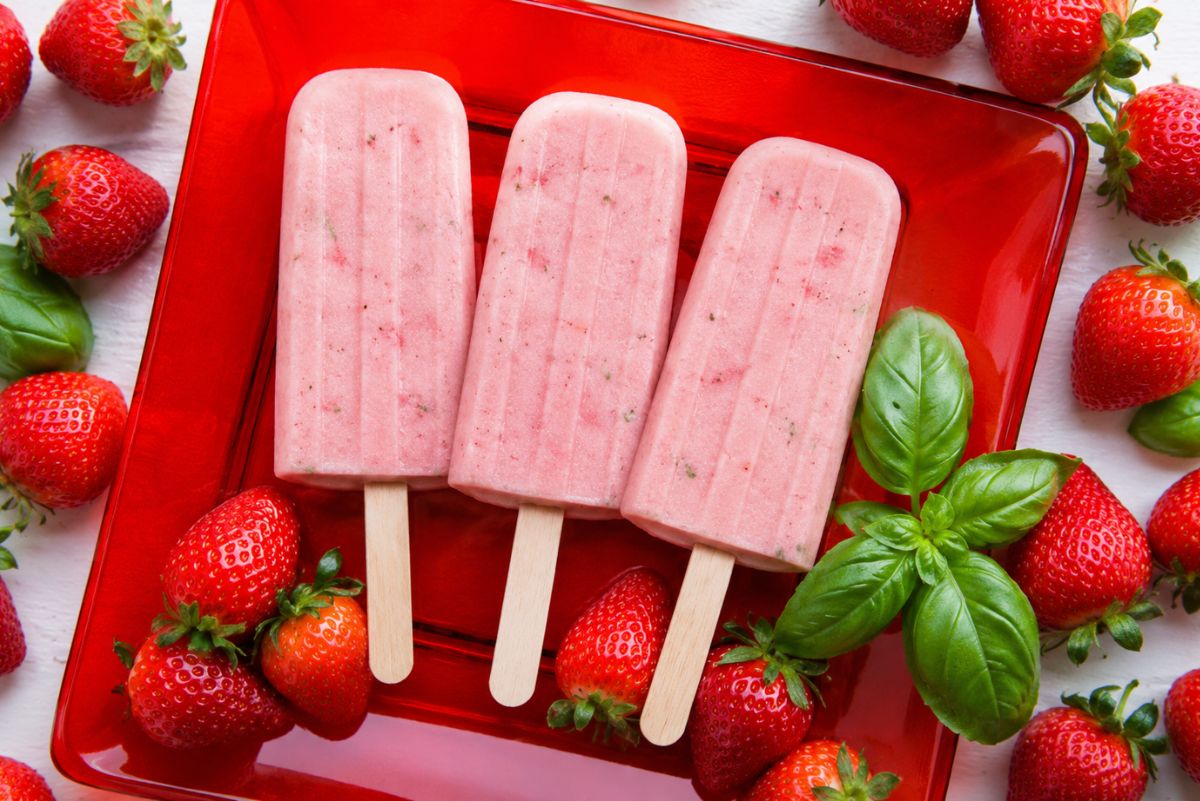 Honey strawberry basil popsicles on a red plate with fresh strawberries and basil leaves on a white wood background.